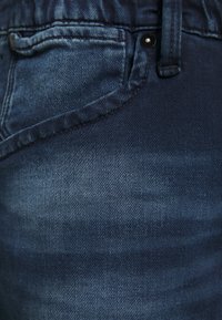 Tumma sininen farkkukangas, jossa on hieman haalistuskuviota, näkyvät ompeleet ja pyöristetty etutasku.