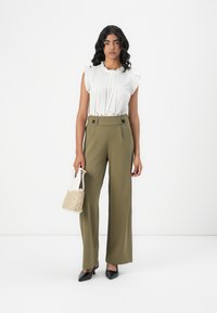 Blusa bianca a balze con dettagli in pizzo abbinata a pantaloni larghi a vita alta di un verde oliva, tacchi a punta neri e una borsa intrecciata.