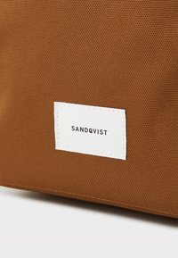 Brązowa torba z teksturowanej tkaniny z białą metką z logo "SANDQVIST". Wytrzymała konstrukcja i prosty design z wykończeniami szwów.