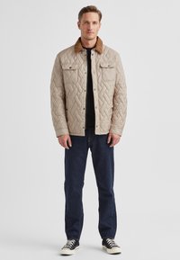 Beige gesteppte Jacke mit braunem Cordkragen, zwei Fronttaschen und einem Metall-Druckknopfverschluss, kombiniert mit dunkelblauen Jeans und Turnschuhen.