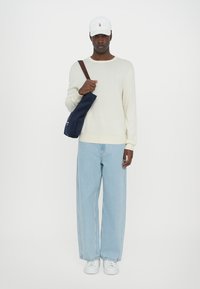 Crème gebreide trui, lichtblauwe wijde jeans, witte sneakers en een marineblauwe tas. Afgerond met een witte baseballcap met een logo.