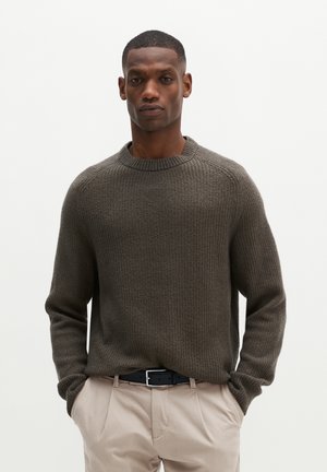 Homme portant un pull en maille gris foncé et un pantalon beige clair, les mains dans les poches, debout devant un fond clair uni.