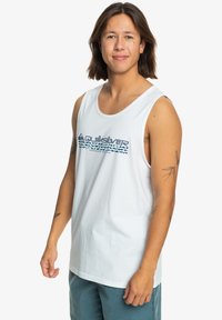 Valkoinen puuvillainen hihaton paita pyöreällä kaula-aukolla, jossa on sininen ja teal Quiksilver-logo keskellä. Sujuva rakenne, rento malli.