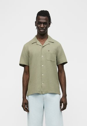 Ung mand med korte dreadlocks iført en lysegrøn kortærmet skjorte med knapper og lyseblå jeans mod en ensfarvet baggrund.