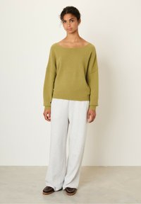 Pull overdimensionné vert olive avec un large col, associé à un pantalon de survêtement gris à jambes larges. Le modèle se tient sur un sol clair, portant des chaussures noires à enfiler.