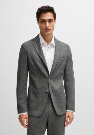 Blazer grigio in lana con vestibilità su misura, revers a punta, due tasche frontali e due bottoni. Indossato sopra una camicia bianca con colletto rigido.