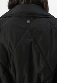 Person wearing a black quilted jacket with diagonal stitching and a small silver “HUGO” logo on the upper back.