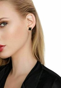 Svarta örhängen med guldfärgade accenter och en vågig design, mot en jämn yta, bärs av en modell.