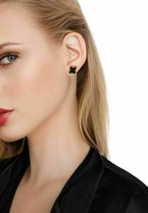Pendientes de stud negros con acentos dorados y un diseño con borde ondulado, sobre una textura suave, llevados en la oreja de un modelo.