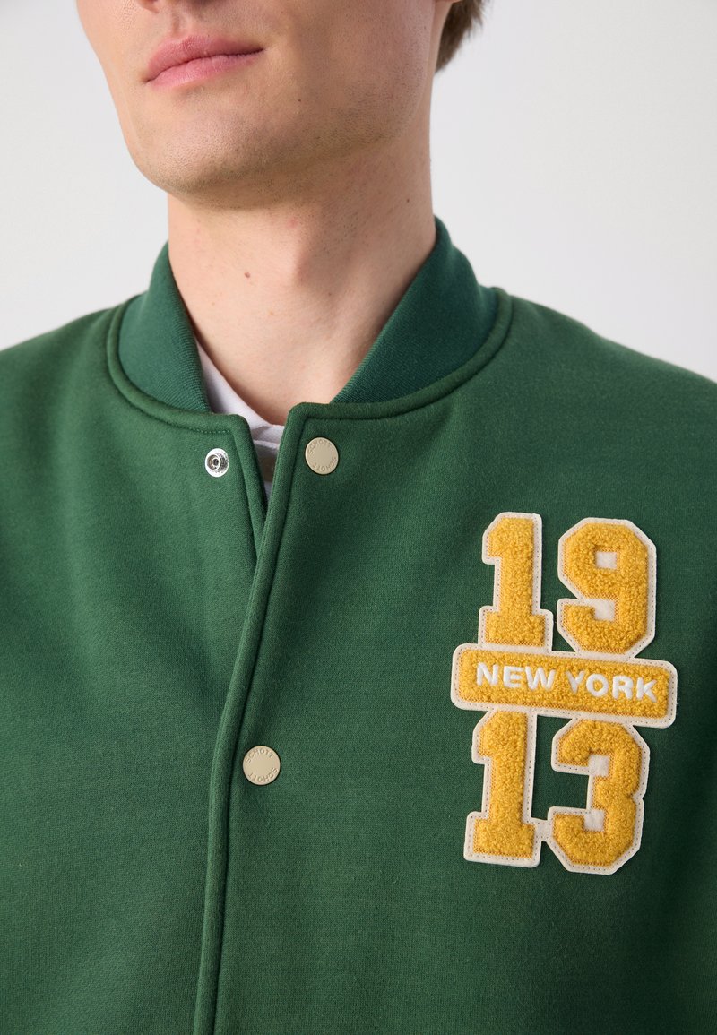 Sweatshirt vert avec un col rond, arborant un patch en feutre jaune portant les chiffres "19" et "13" ainsi que "NEW YORK". Détail de fermeture à pression visible.