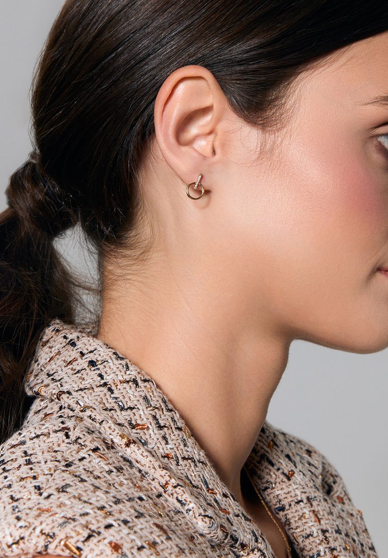Złote okrągłe kolczyki z przezroczystym akcentem, prezentowane na uchu modelki. Modelka ma długie, ciemne włosy i nosi fakturowaną beżową kurtkę.