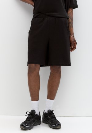 Shorts de sport noirs en tissu texturé, avec une taille élastique et des poches latérales, associés à des baskets noires et des chaussettes blanches mi-mollet.