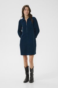Kleid aus dunkelblauem Cord mit Kragen und Reißverschluss vorne, langärmlig und mit geradem Saum. Getragen mit schwarzen Cowboy-Stiefeln.