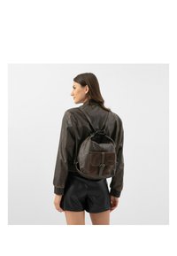 Hnědý kožený batoh s texturovaným povrchem, obsahující přední kapsu, nastavitelné popruhy a decentní logo. Nosený s tmavým kabátem.