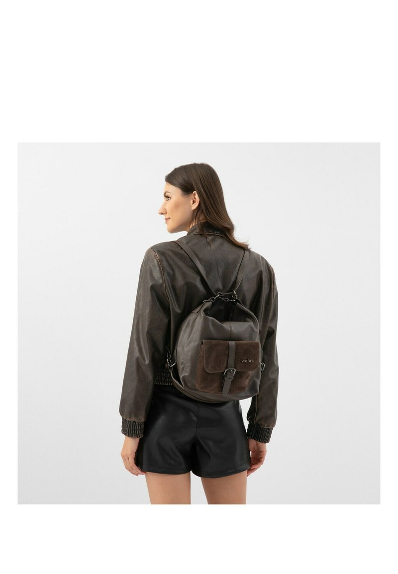 Hnědý kožený batoh s texturovaným povrchem, obsahující přední kapsu, nastavitelné popruhy a decentní logo. Nosený s tmavým kabátem.