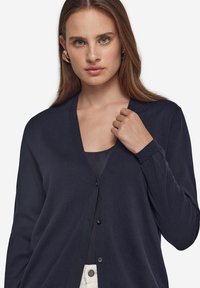 Marinblå cardigan med V-hals, långa ärmar och knäppning. Mjuk textur och strömlinjeformat snitt. Bärs över en mörk topp.