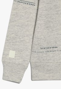 Felpa grigia chiara con una texture morbida, caratterizzata da loghi testuali blu e un piccolo etichetta bianca con un cerchio verde sul polsino.