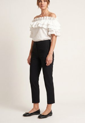 Kvinde iført en hvid bluse med rynkede off-shoulder ærmer, sorte smalle ankellange bukser og sorte balletsko, stående mod en lys baggrund.