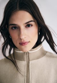 Pull en maille beige à fermeture éclair avec un col montant, présentant un motif texturé et des accents blancs contrastants le long du col et de la fermeture éclair.