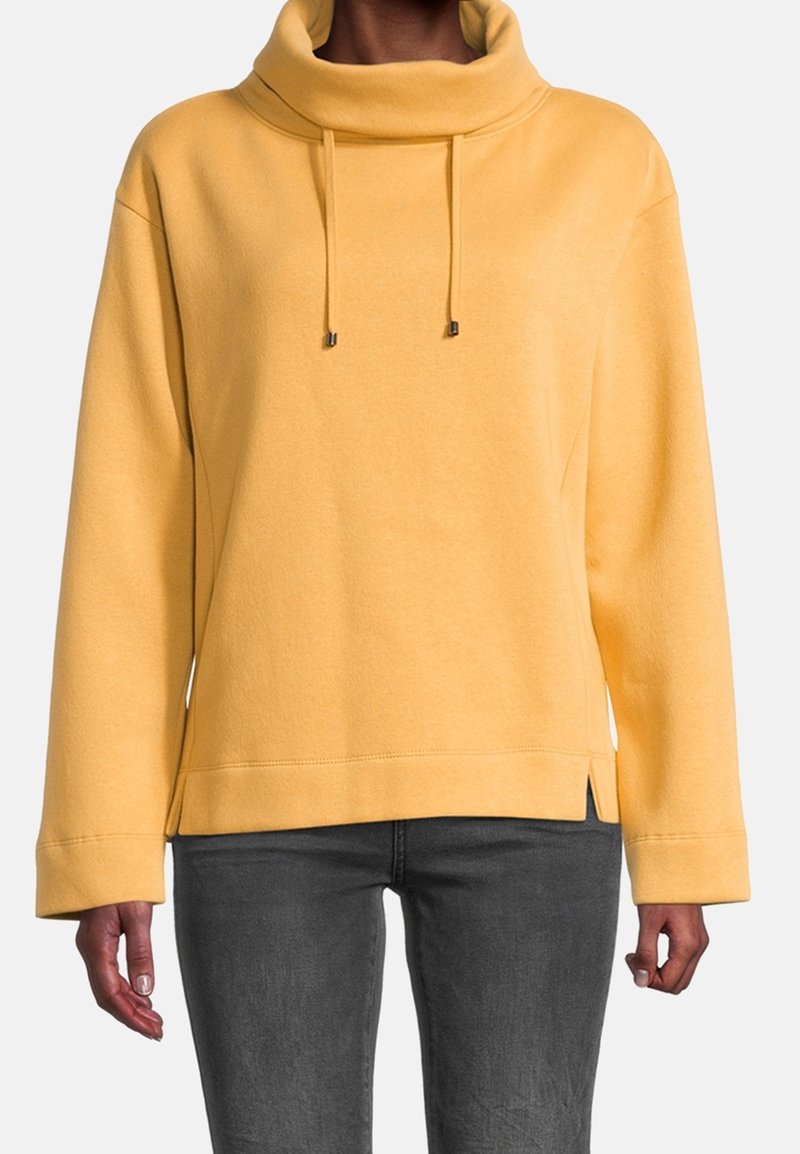 Personne portant un pull jaune à col montant avec cordons et un pantalon ajusté gris foncé, debout devant un fonds uni.