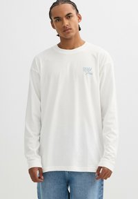 Camiseta blanca de algodón de manga larga con un logo de texto en azul claro en el pecho, combinada con jeans de mezclilla azul claro.