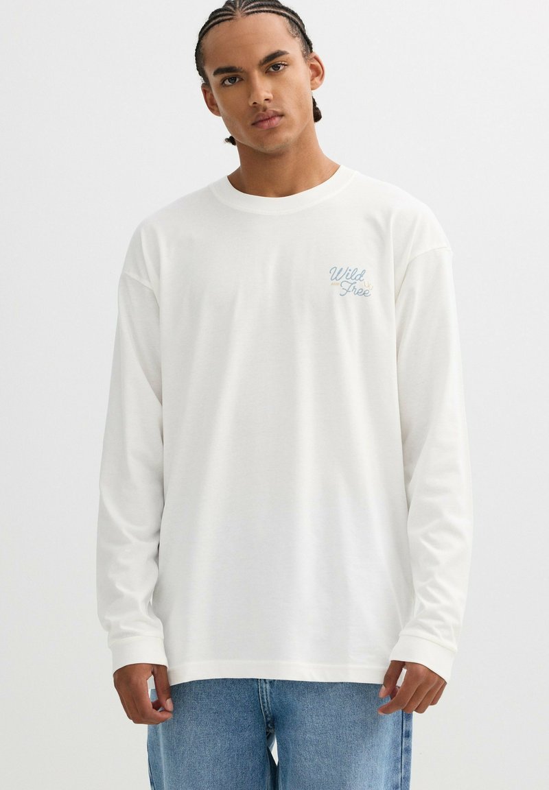 Camiseta blanca de algodón de manga larga con un logo de texto en azul claro en el pecho, combinada con jeans de mezclilla azul claro.