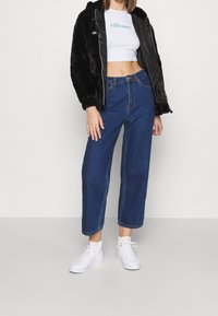 Donna che indossa un top corto bianco, una giacca nera con cappuccio, jeans blu a vita alta e sneakers bianche, in piedi contro uno sfondo semplice.