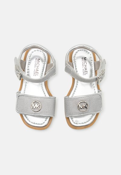 Sandales argentées avec deux brides, l'une comportant un logo en forme de cercle. Le matériau est brillant et texturé, avec une semelle de couleur marron clair. Bride de cheville réglable.