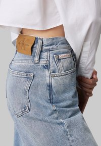 Vaqueros de denim de cintura alta en un tono azul claro, con un parche de cuero marrón y una etiqueta roja de Calvin Klein. Textura desgastada en la tela.
