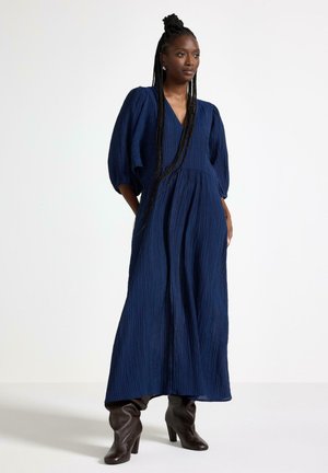 Marineblå kjole med plisseret tekstur, dyb V-udskæring og puffede ærmer. Båret med brune knæhøje støvler med hæl. Enkel, elegant silhuet.