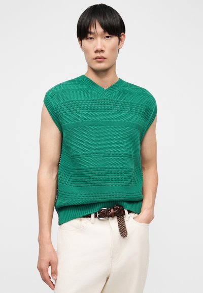 Jeune homme portant un pull-over sans manches vert en maille par-dessus une chemise blanche, avec un pantalon blanc et une ceinture tressée marron, debout devant un fond uni.