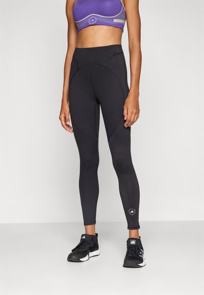 Leggings neri a vita alta con cuciture curve, realizzati in materiale elastico, abbinati a un reggiseno sportivo viola con dettagli in rete e grafiche.