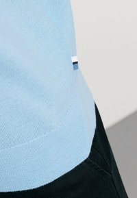 Hellblauer Pullover mit geripptem Muster, ausgestattet mit einem kleinen Seitenschild in Marineblau und Weiß. Über dunklen Hosen getragen, schräger Blick.