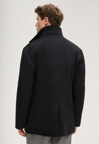 Giacca nera con colletto alto, texture liscia e design strutturato. Presenta un taglio dritto, tasche laterali e dettagli di cuciture minimali.