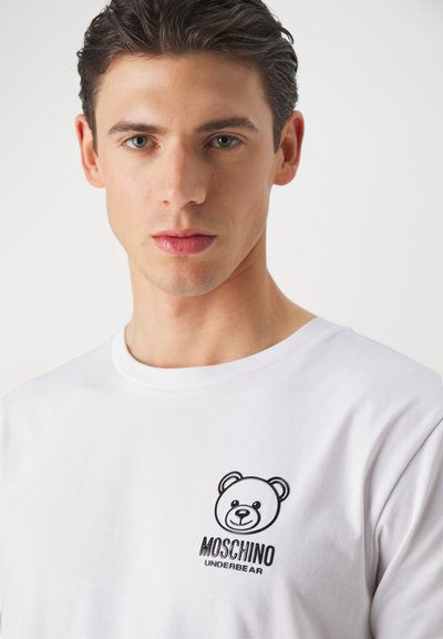 Balta medvilninė marškinėliai su juodo lokio grafika ir "Moschino Underbear" tekstu kairėje krūtinės pusėje. Trumpi rankovės ir apvalus iškirpimas.