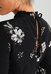 Schwarzes Blumenkleid mit Rüsche am Ausschnitt, offenem Rücken mit Bindeband sowie weißen und rosa Blumenmustern auf dem Stoff.