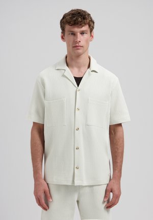 Man draagt een wit wafeltje korte mouwen overhemd met grote borstzakken en bijpassende broek, staand tegen een grijze achtergrond.