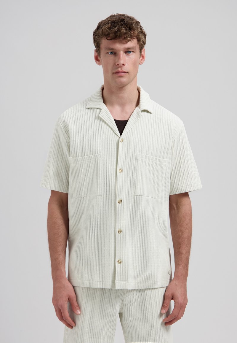 Homme portant une chemise blanche à manches courtes en tissu gaufré avec de grandes poches avant et un pantalon assorti, sur fond gris.