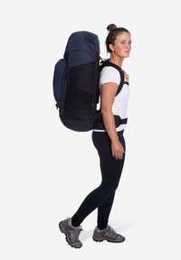 Donkerblauwe en zwarte rugzak met verstelbare banden, bucklesysteem en gestructureerde stof, gedragen door een persoon in zwarte leggings en grijze schoenen.