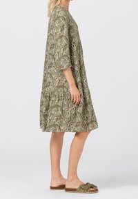 Nainen, joka on pukeutunut väljä, polven pituiseen oliivinvihreään paisley-mekkoon, jossa on kolmen neljänneksen hihat, ja oliivinvihreisiin nauhakorkokenkiin, seisoo sivuttain.