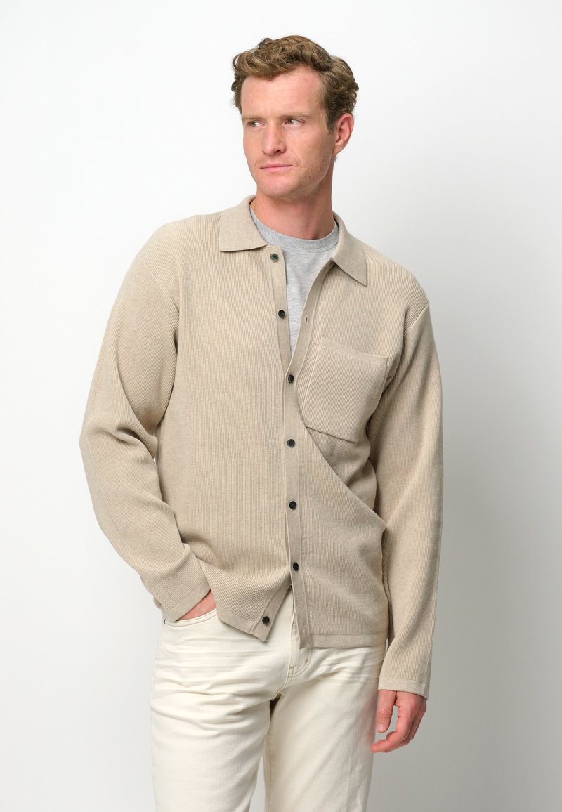 Beige gestrickte Hemdjacke mit kragenartigem Ausschnitt, Knopfverschluss, Brusttasche links und geripptem Muster, getragen über einem grauen T-Shirt und weißen Hosen.