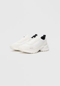 Hvite athletic sko med en kombinasjon av glatte og teksturerte materialer, med et lagdelt design, fremtredende gummisåle og svarte detaljer.