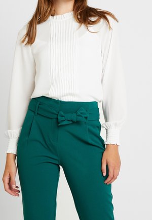 Blusa bianca con frontale plissettato e polsini arricciati abbinata a pantaloni teal a vita alta con un dettaglio a fiocco in vita.