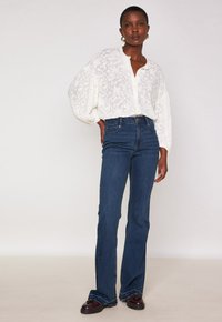 Donna in una trasparente camicia floreale bianca con maniche a sbuffo, jeans scuri a vita alta e svasati, e mocassini scuri, in piedi contro uno sfondo semplice.