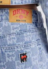 Jeans de mezclilla azul claro con un patrón de logotipo repetido, que cuenta con una etiqueta de cuero color marrón y un parche de logotipo negro y rojo cosido en la parte de atrás.