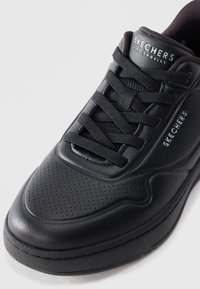 Black leather sneaker with textured surface, perforations near the toe, flat black laces, and "SKECHERS" branding on the side.