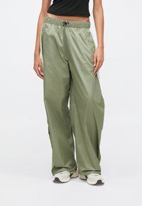 Άτομο που φοράει φαρδιά χακί φόρμα με λευκές και νεονιώδεις κίτρινες λωρίδες στο πλάι, συνδυασμένη με λευκά αθλητικά παπούτσια και μαύρο κοντό τοπ.