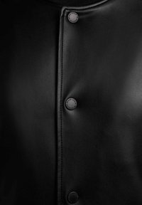 Nahaufnahme eines schwarzen Lederkleidungsstücks mit drei schwarzen Druckknöpfen, auf denen der Markenname entlang einer zentralen Naht geprägt ist.