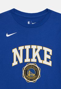 T-shirt Nike bleu avec col rond blanc. Présente un grand texte "NIKE" jaune et un logo circulaire "Golden State Warriors" au centre.