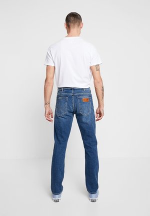 Homme portant un t-shirt blanc et un jean en denim bleu vu de dos, debout devant un fond clair uni.